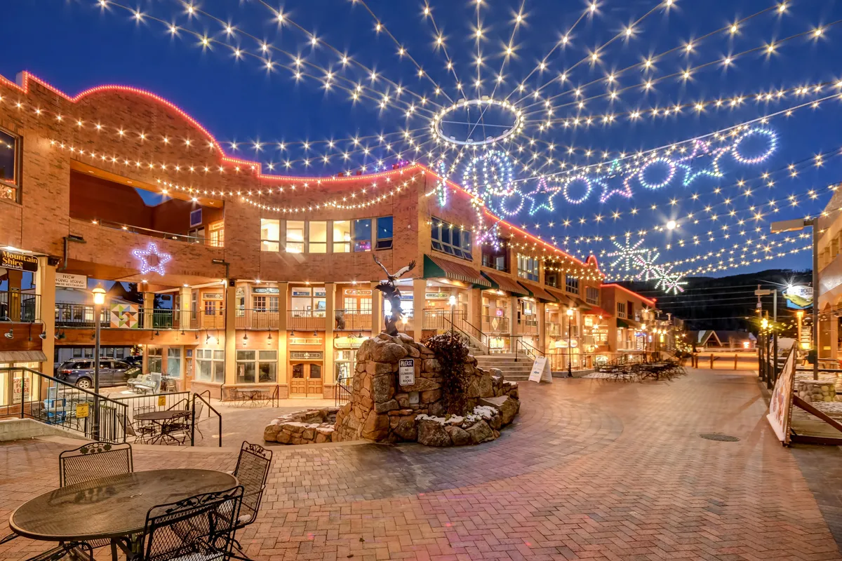 An inviting exterior view of a lively downtown scene with decorative string lights, a brick pathway, central stone feature, and welcoming storefronts. The image exudes a sense of celebration, evening strolls, and holiday events.