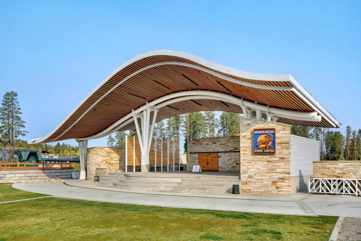 An exterior scene featuring a curved roof structure and stone walls forming a stage area, surrounded by open space ideal for gatherings. The setting is vibrant, suggesting community events, concerts, and outdoor celebrations, with a festive atmosphere.