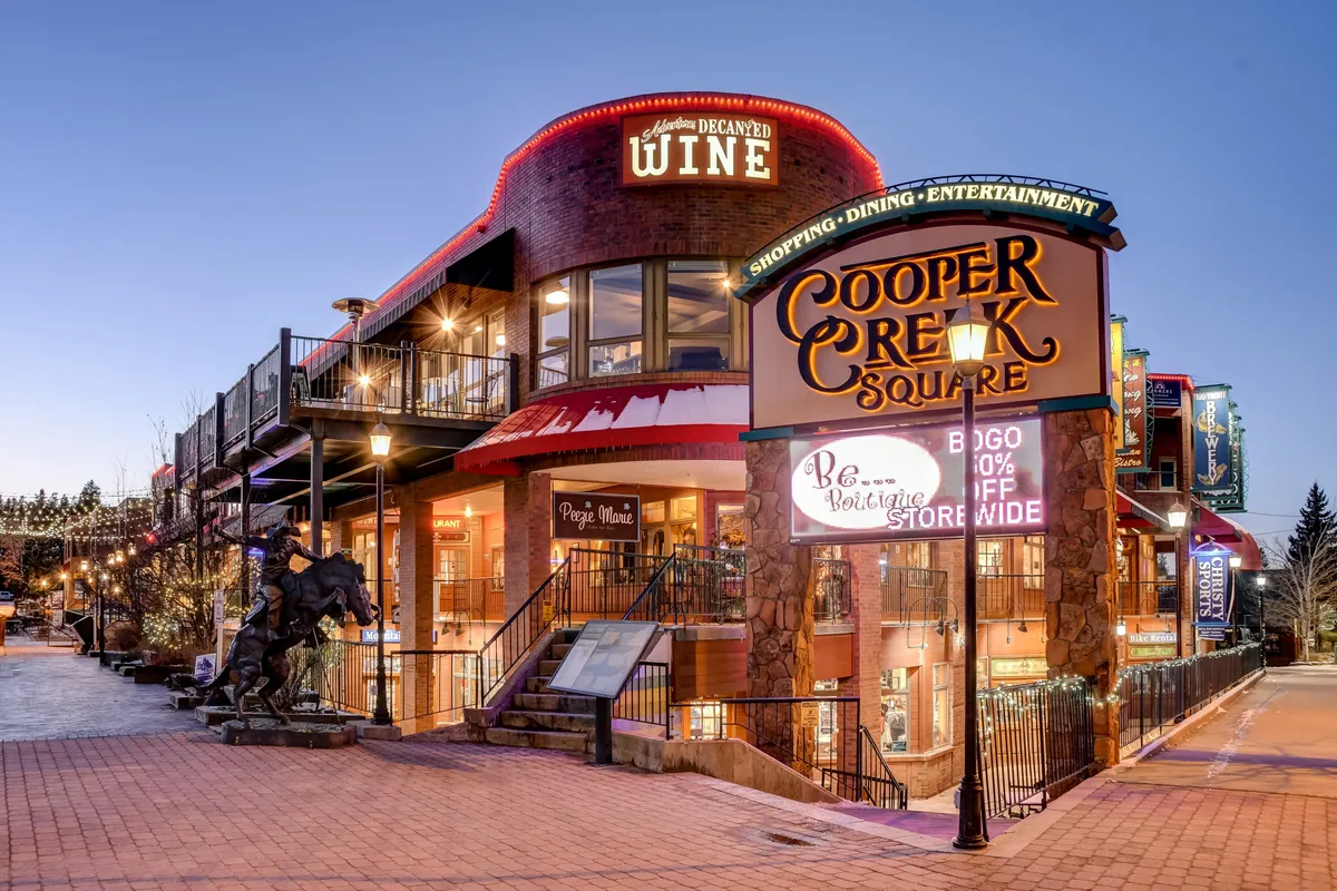 A lively street scene showing Cooper Creek Square, with signs for dining and wine shops, an outdoor seating area, and a welcoming walkway statue. The area is bustling with activity, highlighting the proximity to boutique shopping and eclectic dining options referenced in the section.