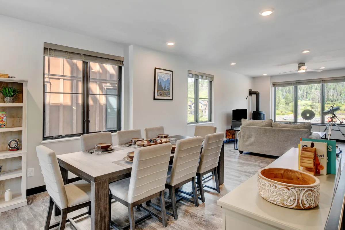 This image showcases an open-plan layout encompassing a dining table and adjacent living room seating, all bathed in natural light from large windows. The windows provide a glimpse of the landscape outside, reinforcing the sense of spaciousness and connection to the environment. The arrangement invites gatherings, family meals, and relaxed socializing, perfectly embodying the heart of the home.