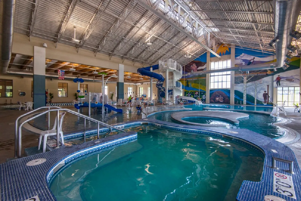 The image shows a vibrant indoor recreation center with a swimming pool, colorful water slides, a cheerful mural, and children playing. Lounge chairs line the pool area, creating a fun and energetic atmosphere perfect for families.