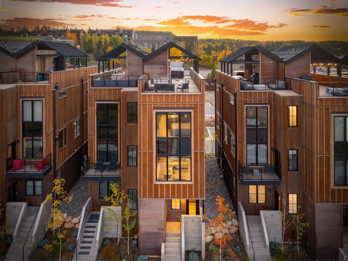 The hero image showcases a modern single-family home with striking wooden facades and expansive windows, featuring inviting rooftop terraces. Guests are seen gathering on the rooftops, enjoying vibrant sunset views, while others stroll along the scenic pathways in a lively yet tranquil neighborhood setting.