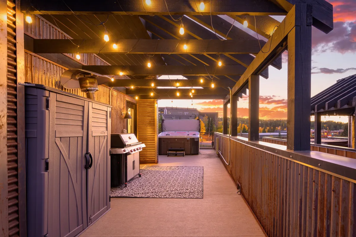 This image features a cozy rooftop patio scene with string lights illuminating the space, a bubbling hot tub, a BBQ grill, and a rug. The ambiance is warm and welcoming, with seating areas designed for relaxation and social gatherings, set against the backdrop of a mountain sunset.