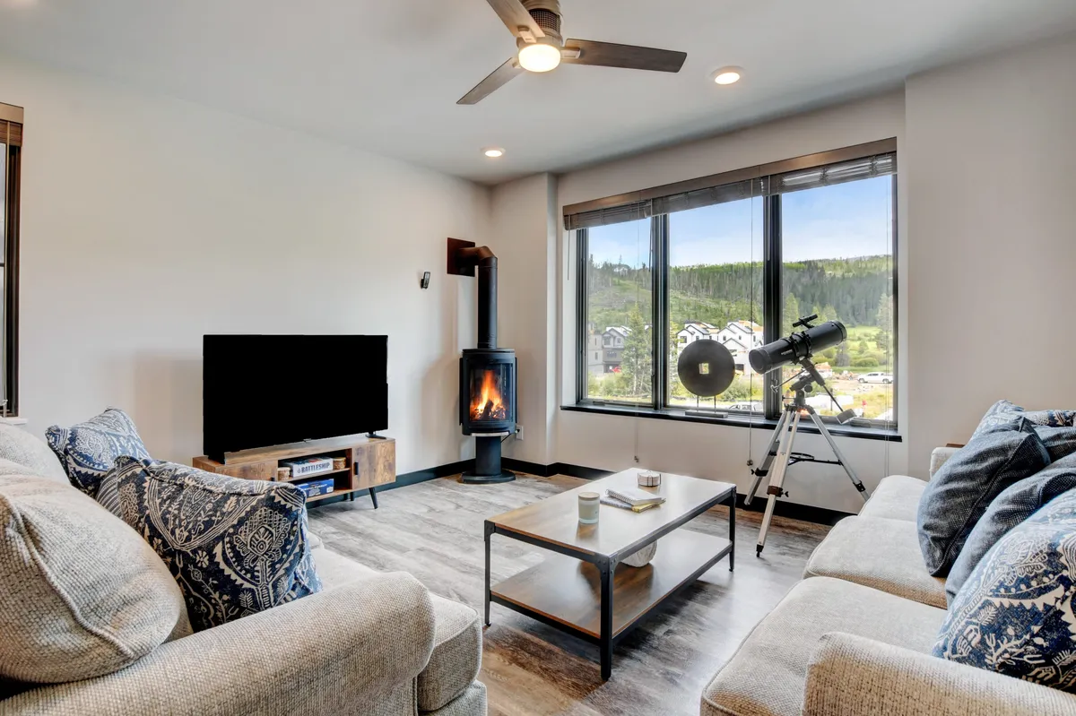 A cozy living room featuring a plush sofa, a large flat-screen TV, a modern fireplace, and a telescope positioned by expansive windows. Natural light floods the space, and the setup encourages relaxation, family movie nights, or stargazing.
