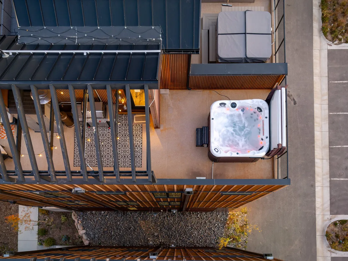 A tranquil rooftop setting with a hot tub, outdoor seating area, string lights, and wooden panels. The space feels intimate and serene, perfect for soaking in the hot tub while enjoying sunset views or gathering for evening conversations under the twinkle of lights.