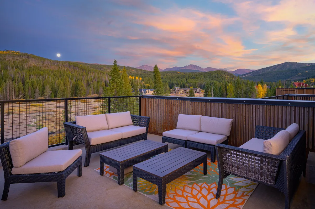 A serene exterior setup featuring a comfortable seating arrangement that faces a breathtaking mountain view. The sky is painted with the colors of sunset, creating a tranquil atmosphere perfect for winding down. Plush outdoor seating invites groups to gather and unwind, while the setting sun highlights the natural beauty surrounding the property.